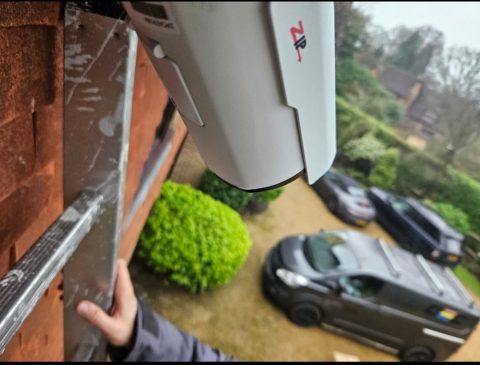 A CCTV camera being fixed to an outside wall, looking down over a driveway